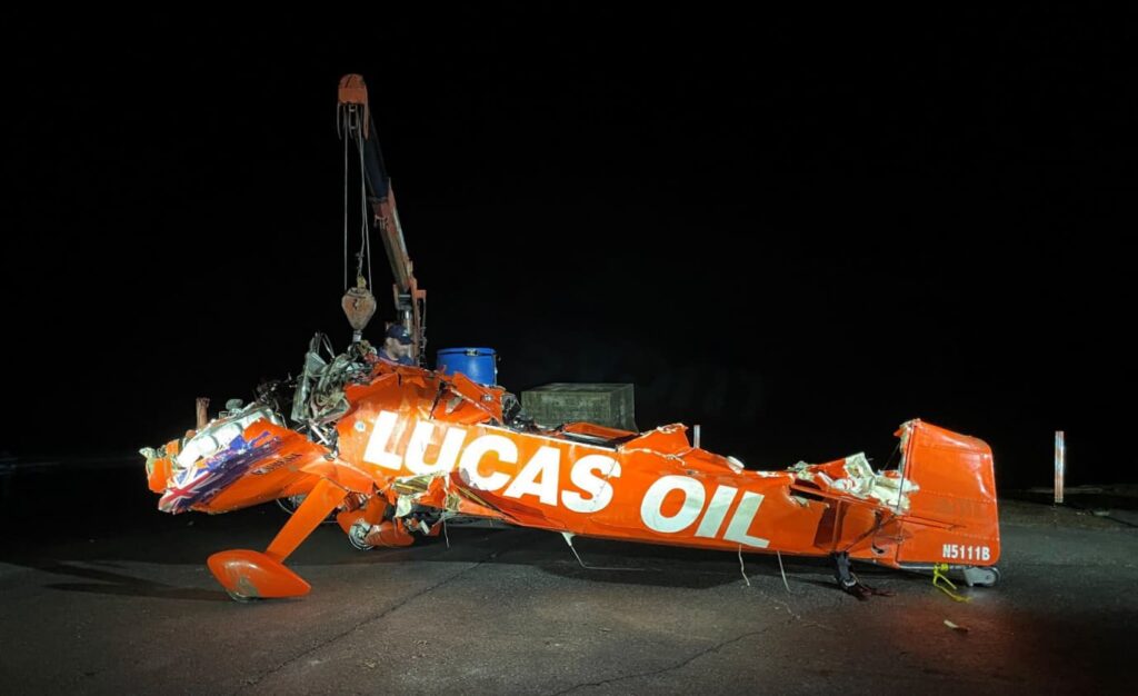 Crushed red Aviat S-1-11B aircraft being lifted by a crane at night. The aircraft bears the “LUCAS OIL” logo and shows extensive structural damage, with the fuselage visibly deformed and missing components.